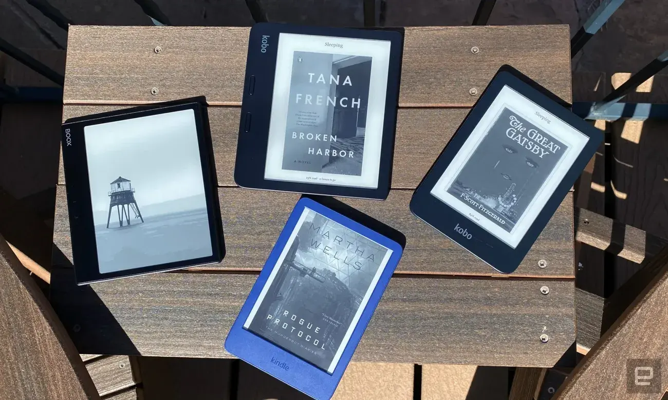Four ereaders are arranged on a dark brown wooden table outside. There are Boox, Kindle and Kobo devices showing the covers of different novels from Tana French, F. Scott Fitzgerald and Martha Wells.