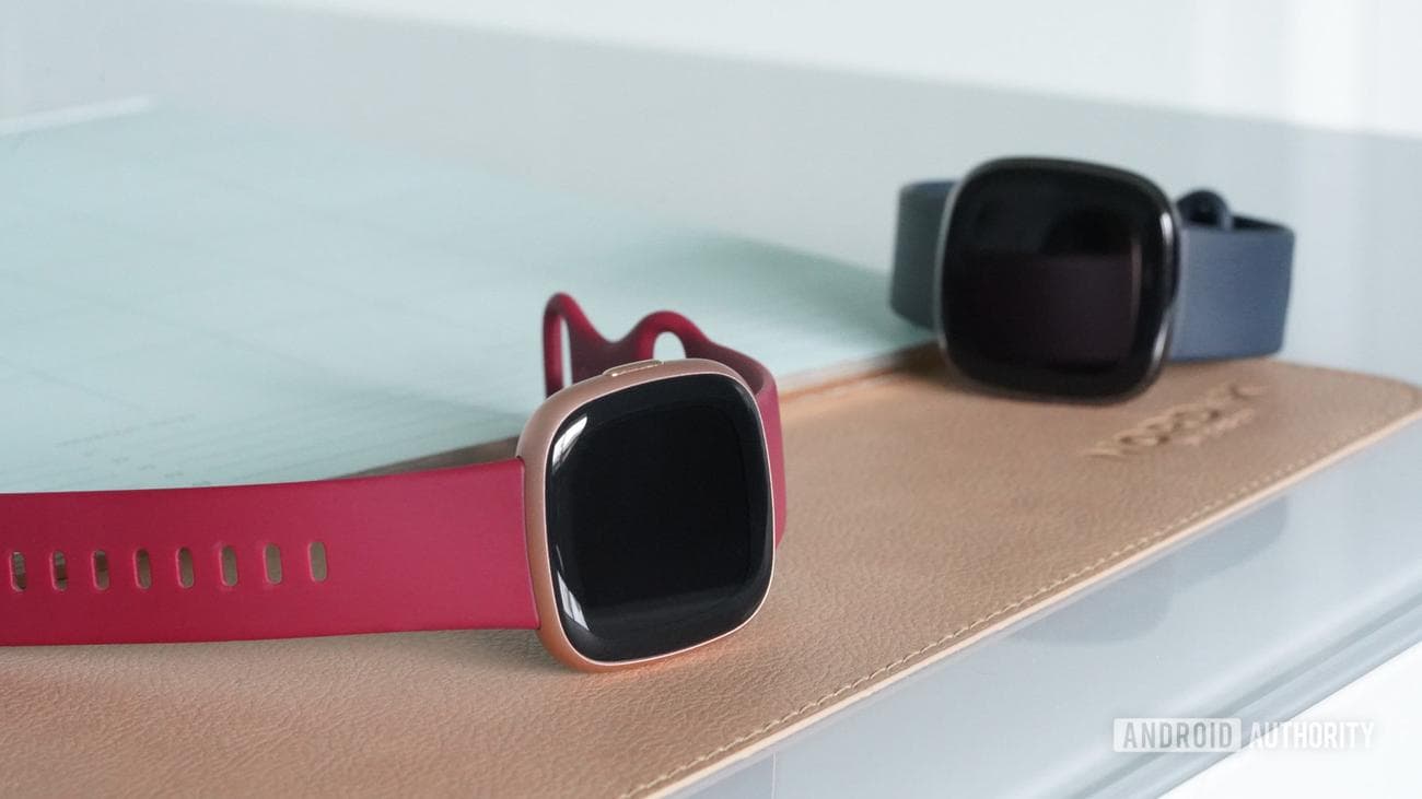 Ένα Fitbit Versa 4 στηρίζεται σε ένα γραφείο δίπλα σε ένα Fitbit Sense.