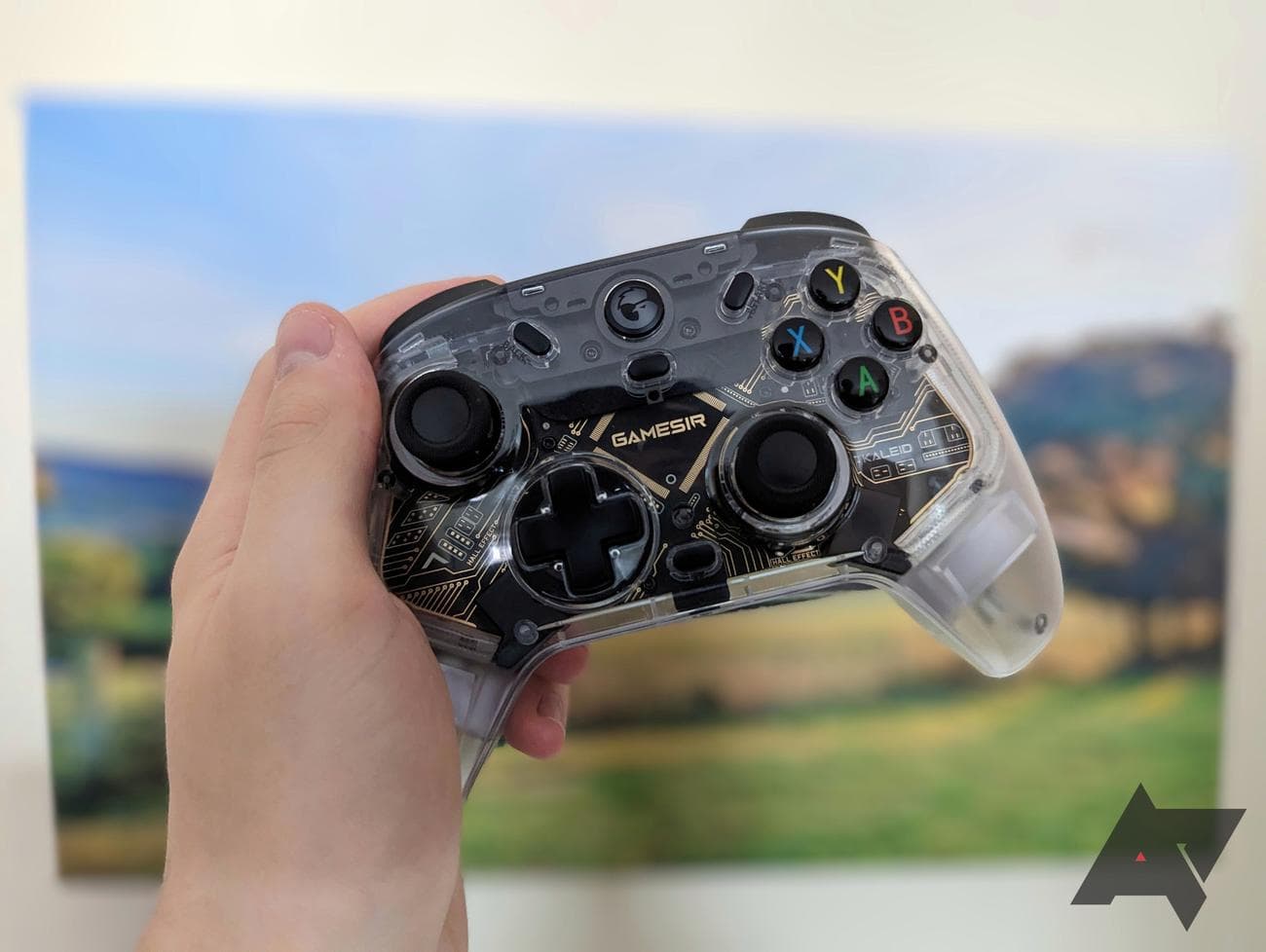 αριστερό χέρι που κρατά καθαρό χειριστήριο παιχνιδιού μπροστά από την εικόνα τοπίου