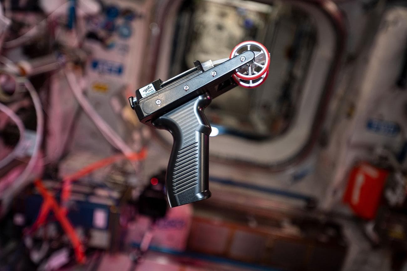 Small hot glue gun-like printer with red wheels floats in zero gravity on the ISS