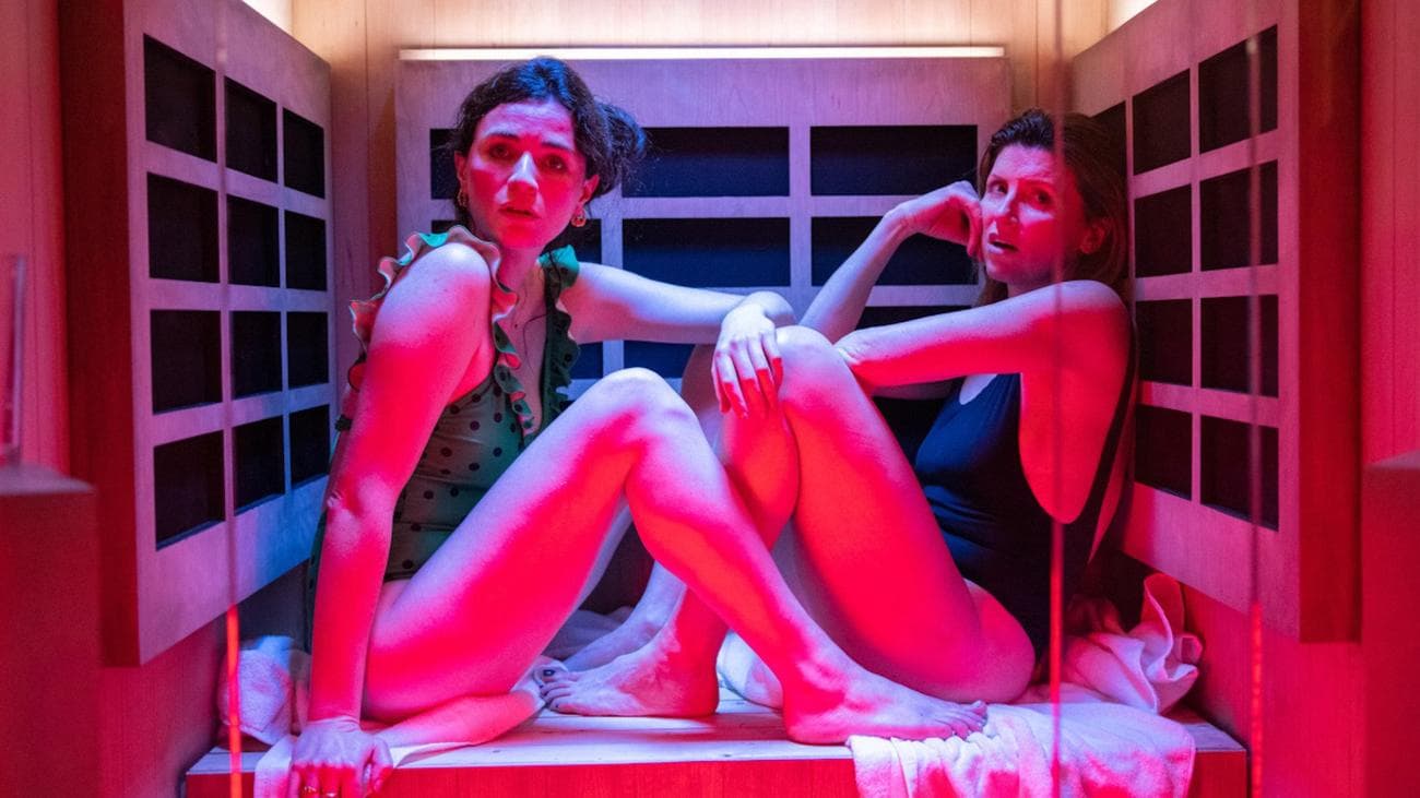 Two women in swimsuits sit in a pink-lit sauna space.