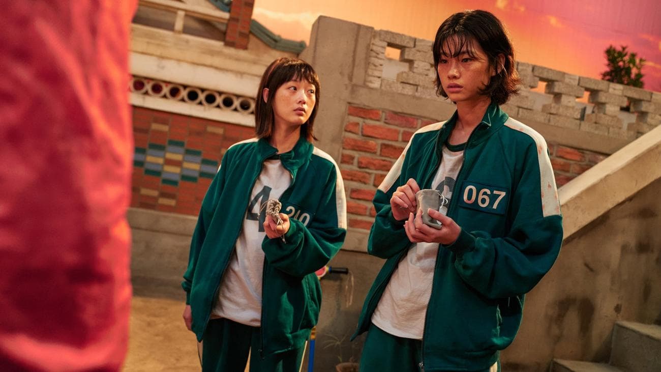 Two women in green jumpsuits stand on a game set.