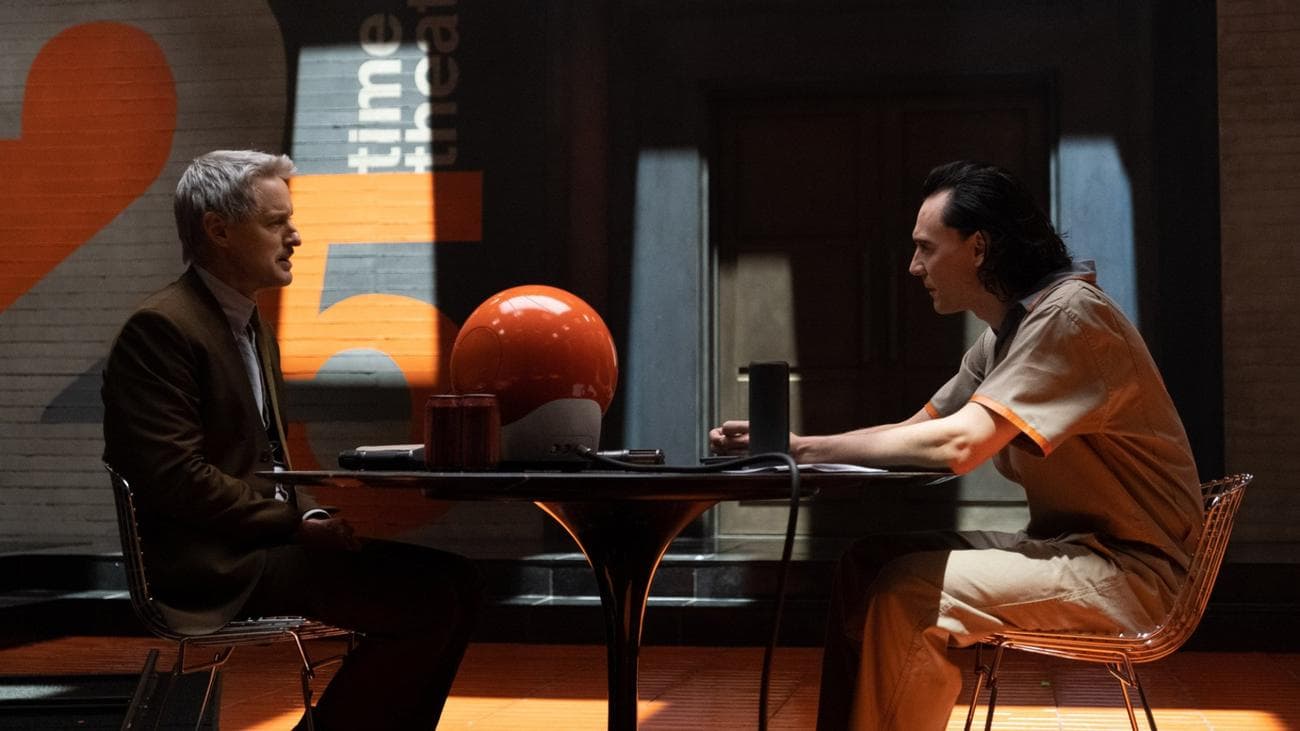 A man in a suit sits across from a man in a prison uniform with a circular orange computer between them.