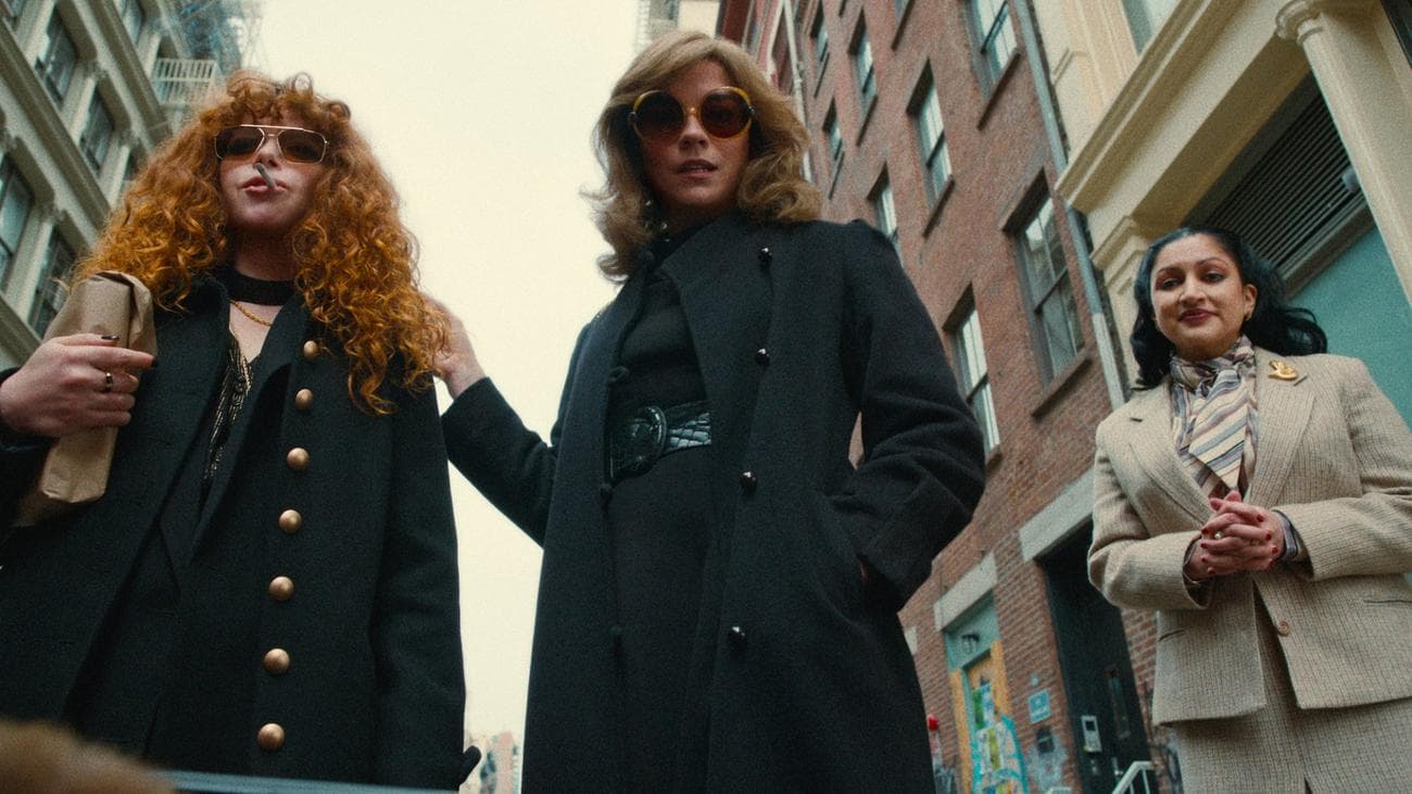 Three women look into a car boot, wearing suits and coats and sunglasses.