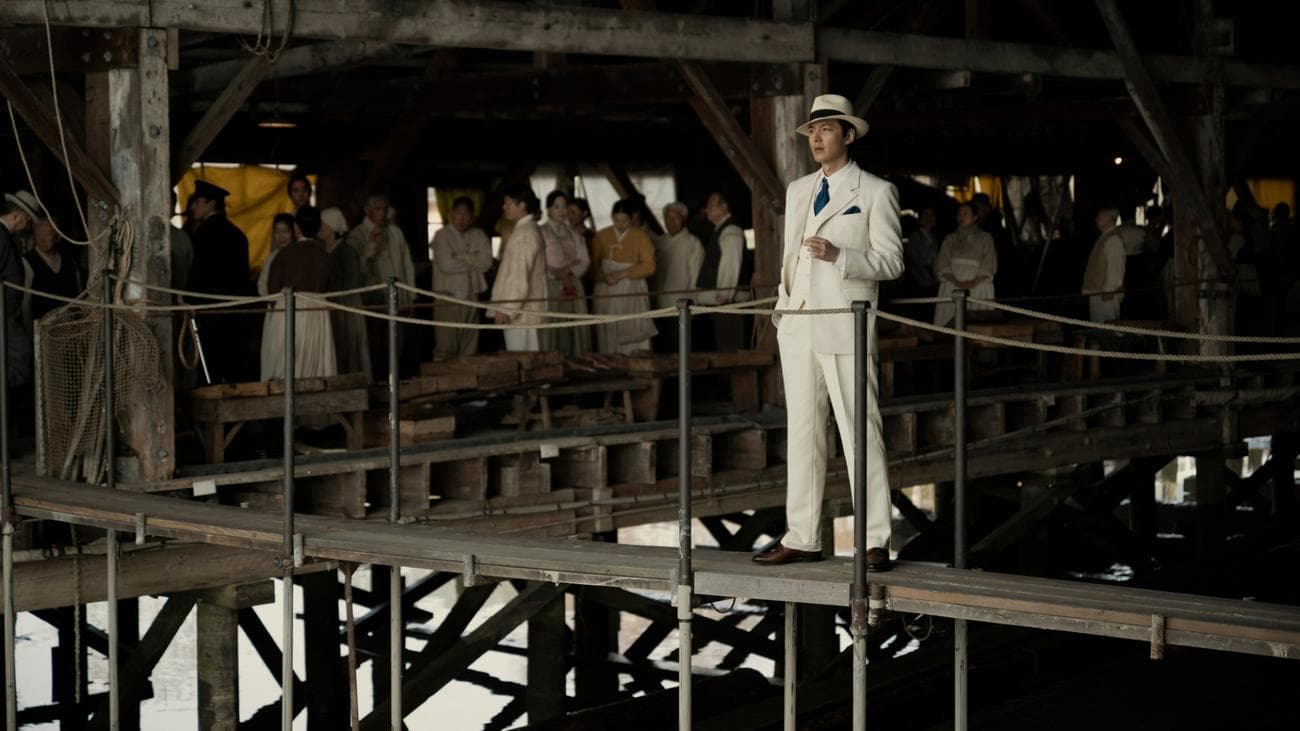A man in a white suit and hat stands on a wooden platform in front of a crowd.