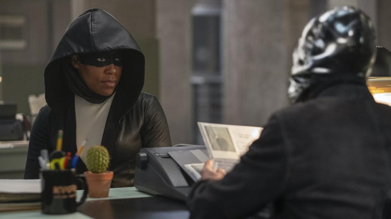 A woman in an eye mask and hood sits at a desk across from a man in a full head mask.