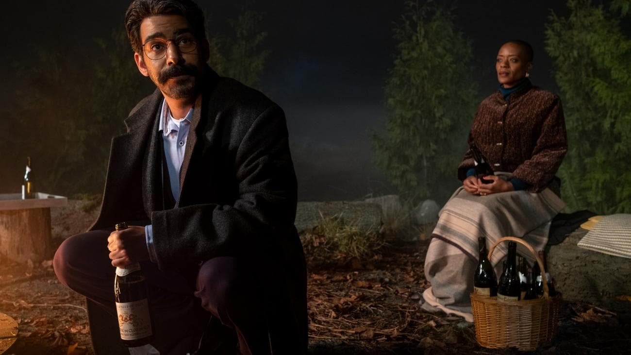 A man sits next to an outdoor fire with a bottle of wine as a woman sits with a glass behind him.