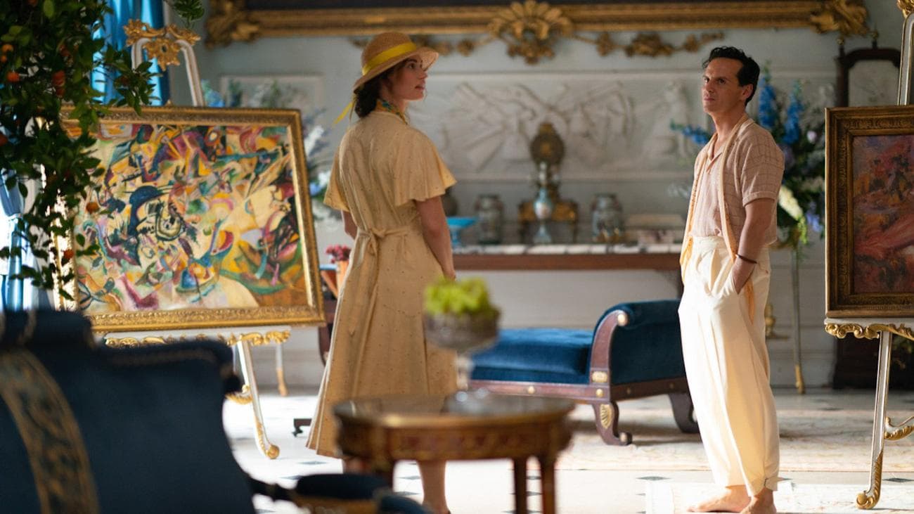 A man and a woman stand in a fancy home next to a painting in a gold frame.