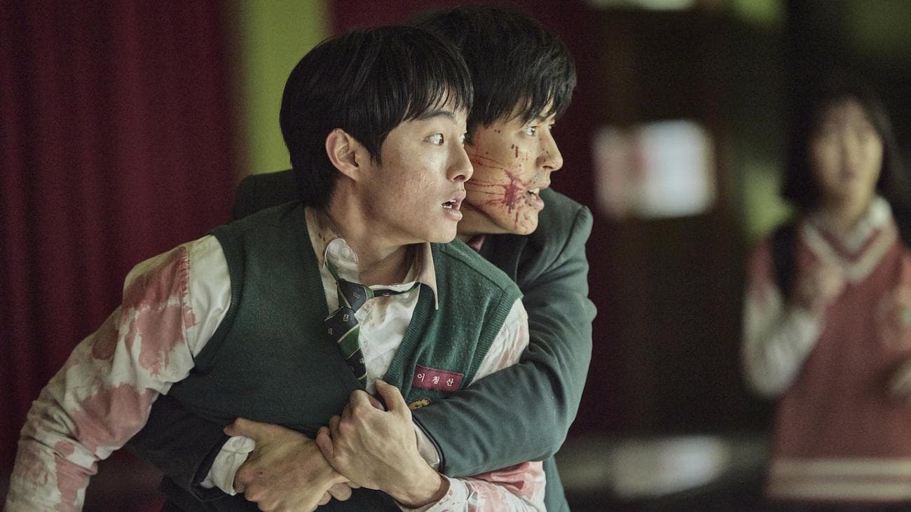 Two school students covered in blood look horrified and hold onto each other.