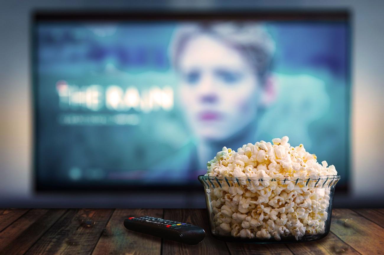A small bowl of popcorn and a television remote control site on a wooden table. A television with a screen is in the background.