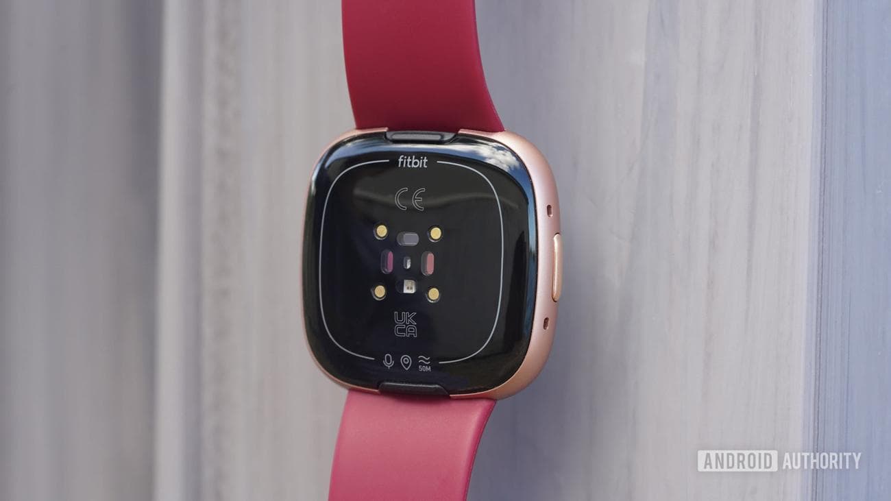 Ένα Fitbit Versa 4 κρέμεται μπροστά από μια γκρίζα επιφάνεια, εμφανίζοντας τους αισθητήρες της συσκευής.