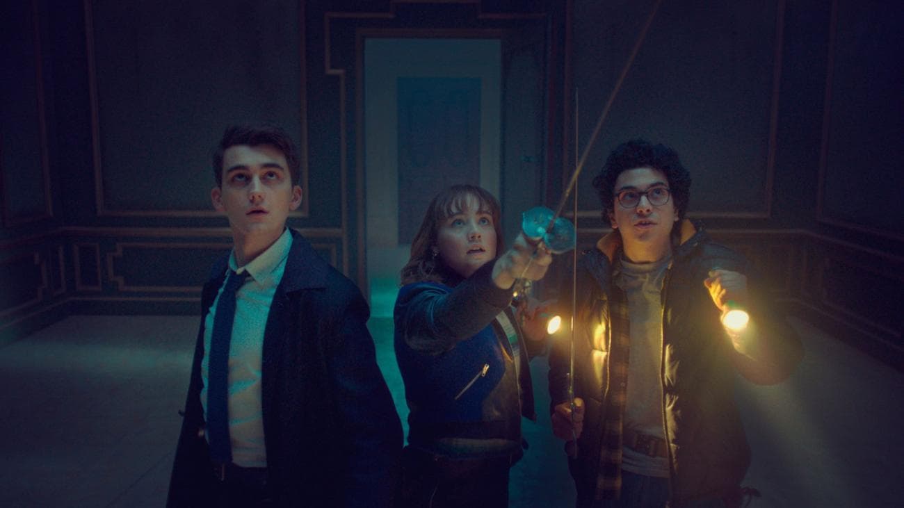 Three teens stand in a dark house with torches and swords in the show "Lockwood and Co"
