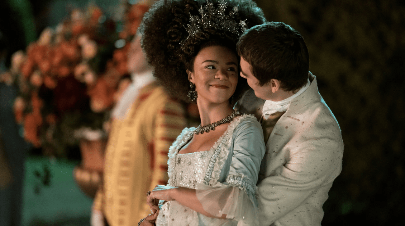 A man in regal dress embraces a woman in regal dress, and the two smile at each other.