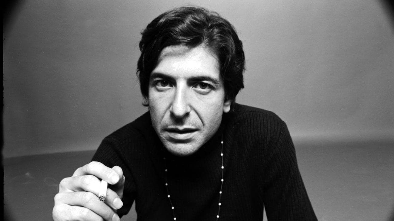 Portrait of Leonard Cohen dressed in black, as he holds a cigarette in one hand.