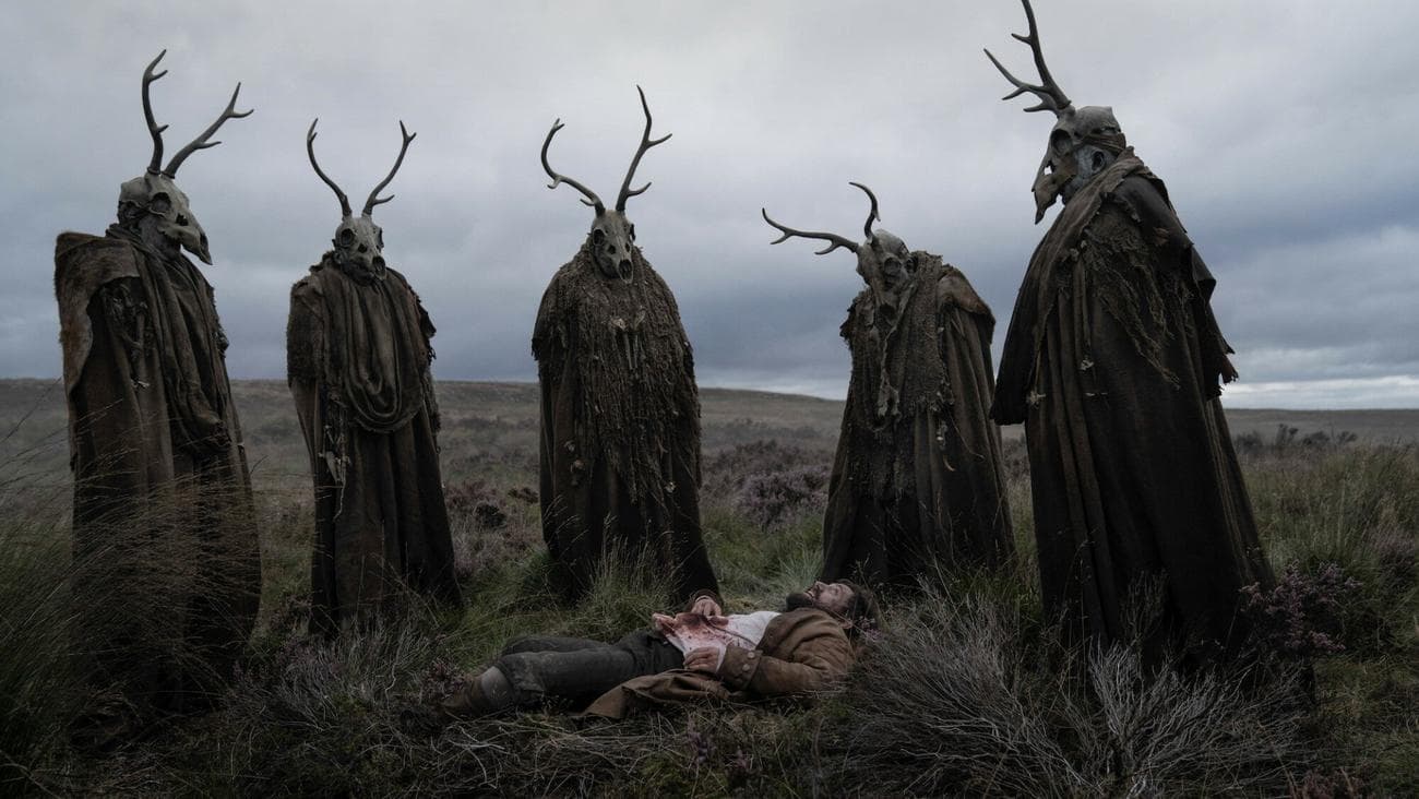 Five figures in cloaks wearing deer skulls as masks gather around a man lying on his back with blood on his shirt.