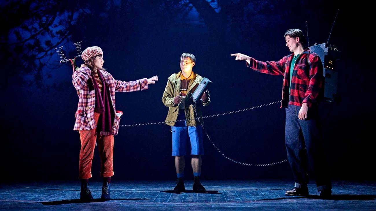 Σε "Stranger Things: The First Shadow" τρεις έφηβοι ενώνονται με μια μεγάλη συσκευή ραδιοφώνου, που δείχνει ο ένας στον άλλο.