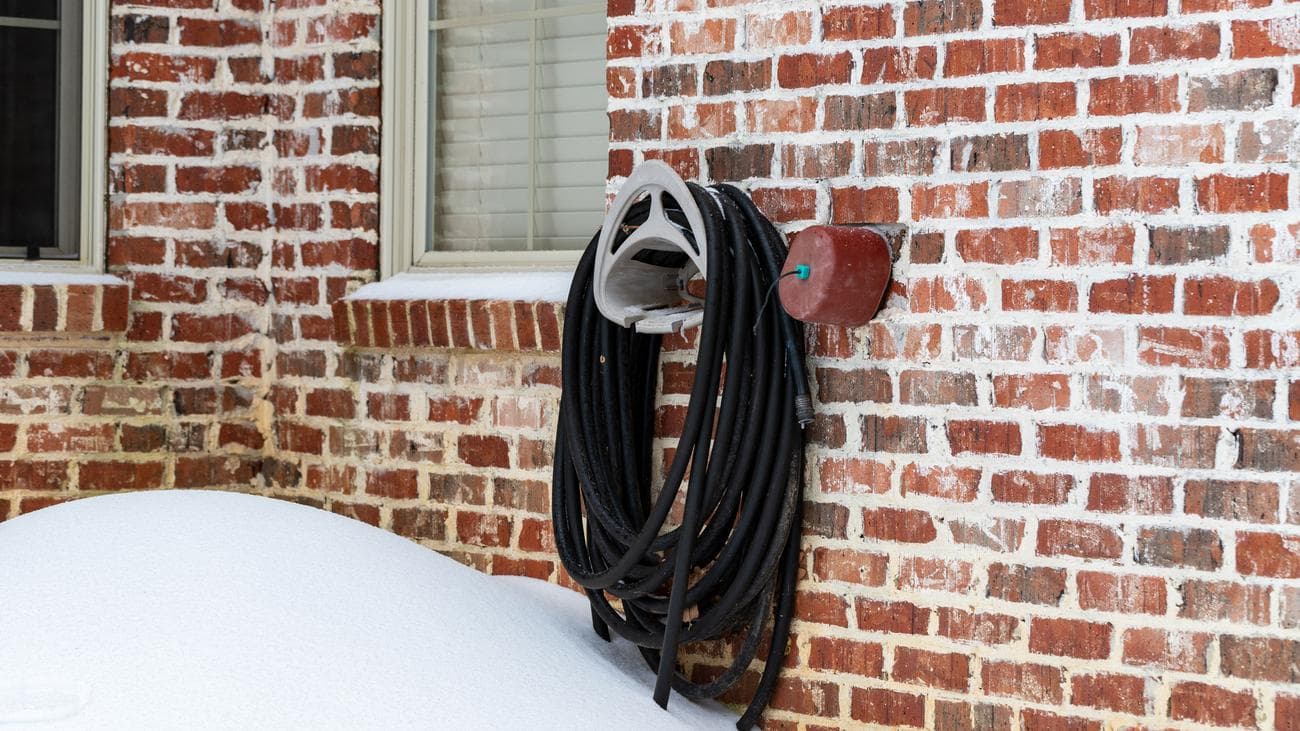 Μια βρύση κήπου με ένα κάλυμμα πάνω της δίπλα σε έναν εύκαμπτο σωλήνα που είναι αποθηκευμένος στον τοίχο