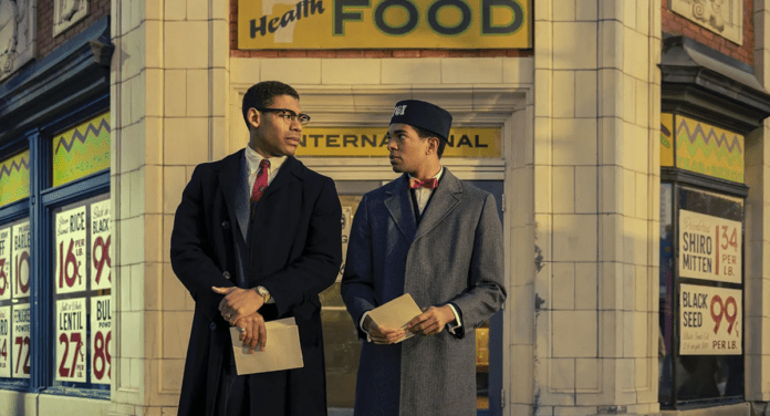 Aaron Pierre star of Genius: MLK/X, wearing a smart suit and red tie, stands in front of a vintage American building 