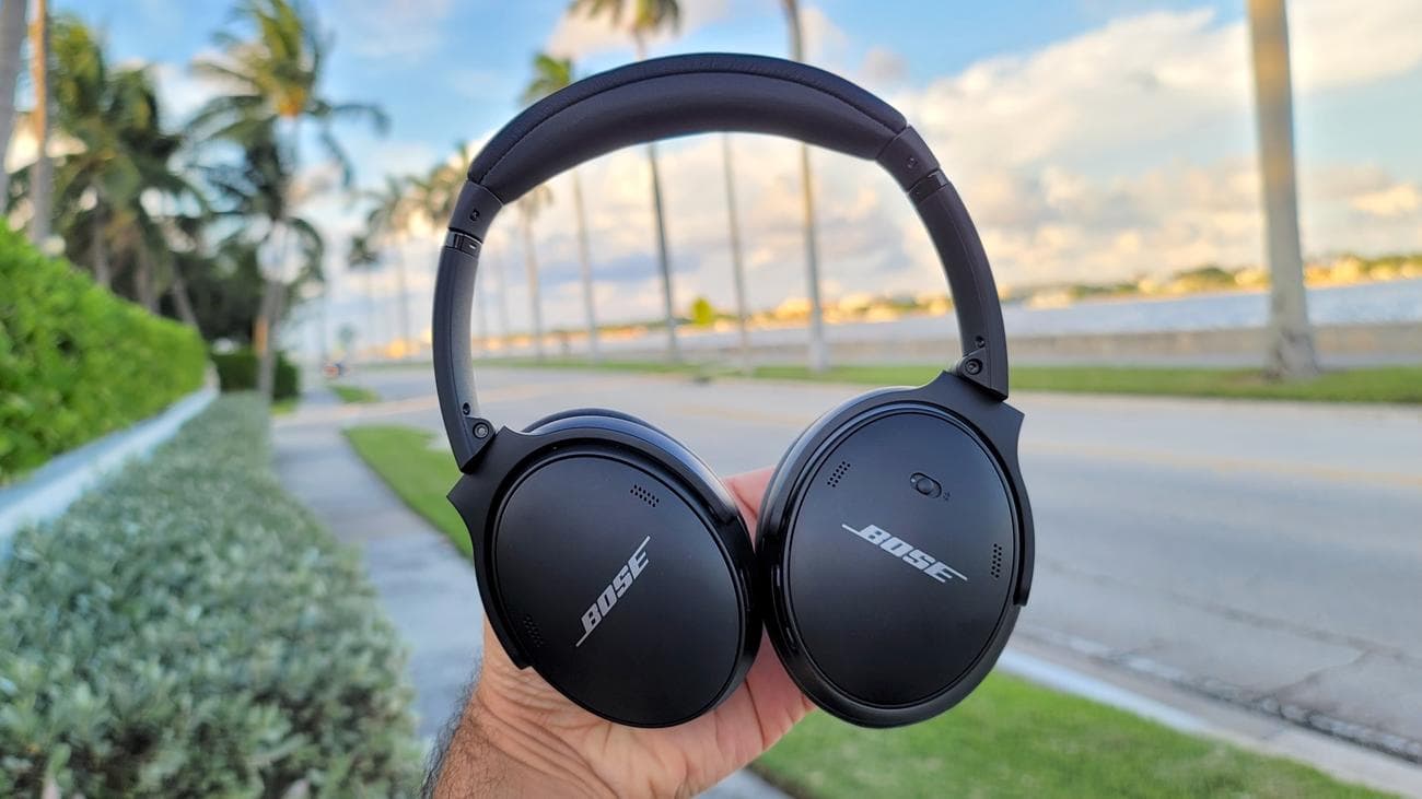 Τα ακουστικά Bose QuietComfort 45 κρατιούνται ψηλά με φόντο έναν παραλιακό δρόμο με φοίνικες