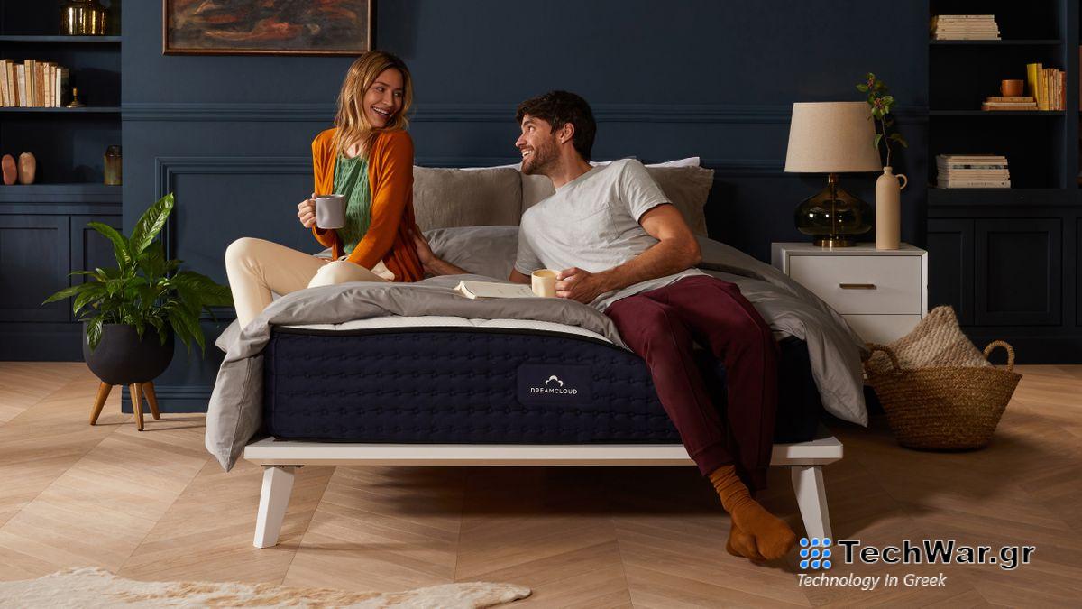 The DreamCloud Hybrid Mattress photographed on a wooden bedframe placed in a dark blue bedroom and with a couple laughing and sitting on the mattress