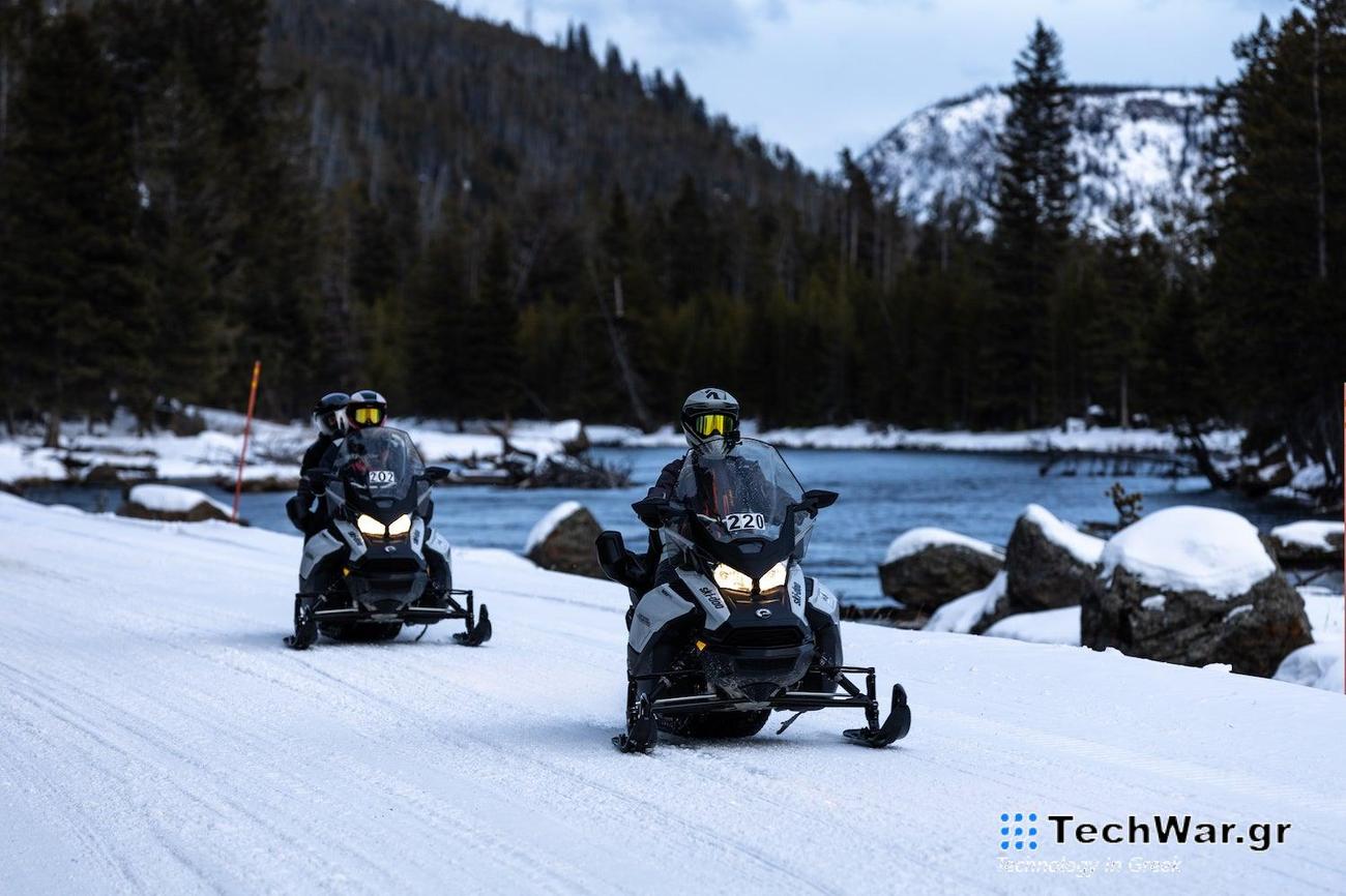 Two snowmobiles ride on the snow next to a river with a mountain and forest in the background